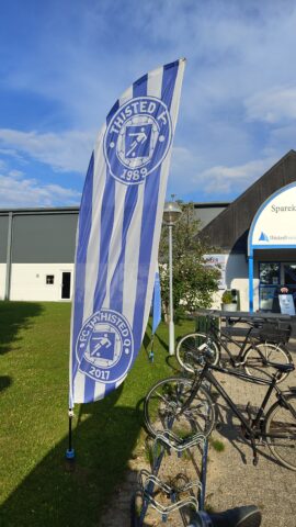 Fahne des Thisted FC mit blau-weißen Streifen und Vereinslogo, aufgestellt vor dem Stadion bei sonnigem Wetter.