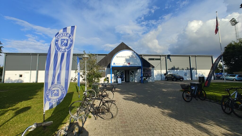 Eingang zur Sparekassen Thy Arena, Heimatstadion von Thisted FC, umgeben von Fahnen und Fahrrädern.