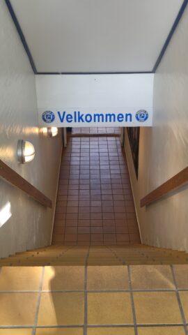 Treppenhaus mit dem Schriftzug 'Velkommen' an der Wand, das Licht fällt von oben herab.