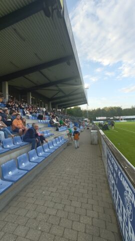 Blick auf die Haupttribüne in der Sparekassen Thy Arena während eines Fußballspiels. Einige Zuschauer sind auf den Sitzen verteilt, der Platz ist teilweise leer. Himmel ist blau mit einigen Wolken.